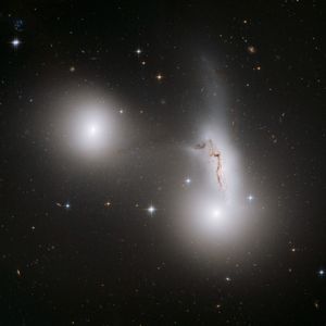 Two glowing white spheres, one on the left above center and one just lower-right of center. Above the right galaxy is a smear of white stars with a streak of brown gas in it.